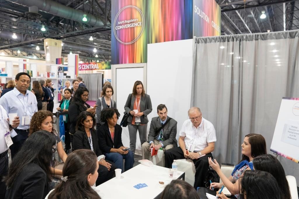 A group of regulatory professionals listen to a speaker in an informal gathering on the show floor of the 2019 RAPS Convention.